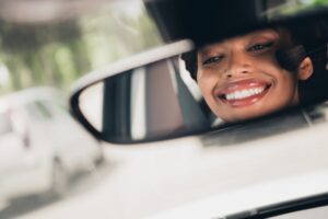 Smiling young woman with radiant smile captured in rearview mirror of a car during sunny day, showcasing joy and beauty